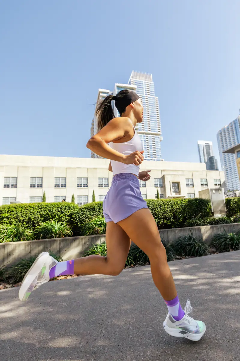 Lizzie running along a city sidewalk in a white tank, lavender shorts and lavender Balega socks, with downtown towers behind her.