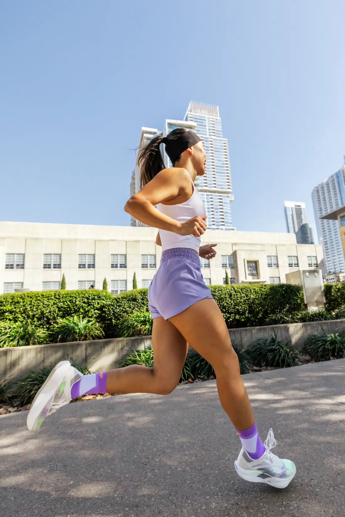 Lizzie running along a city sidewalk in a white tank, lavender shorts and lavender socks, with downtown towers behind her.